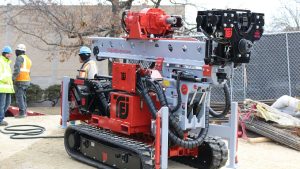 TEI TD100 electric limited‑access track drill rig from National Piling Products set up on a commercial jobsite, ready for micropile and anchor drilling while crew members prepare nearby.