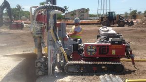 TEI TD75 electric track drill rig from National Piling Products installing micropiles on a commercial jobsite while an operator controls the mast next to a deep excavation.