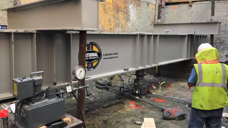 National Piling Products 800‑kip steel test beam assembly set up in a shored excavation, with hydraulic jacks and gauges applying load to foundation piles while a technician monitors the test.