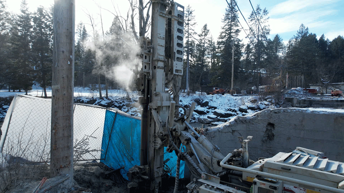 Comacchio MC 14 fully articulated microdrilling rig installing anchors or micropiles beside a winter riverbank, steam rising from the mast while drilling next to a shored excavation.