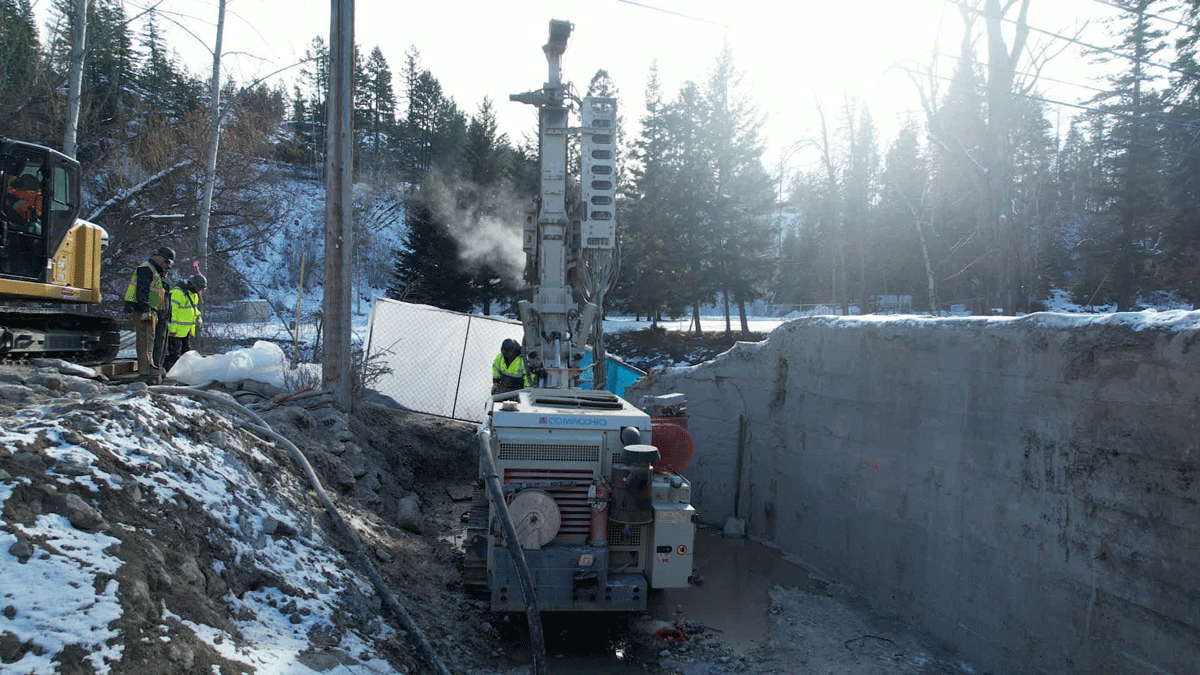 Comacchio MC 14 fully articulated microdrilling rig working in a narrow, shored winter excavation beside a river, with crew members and an excavator observing from above.