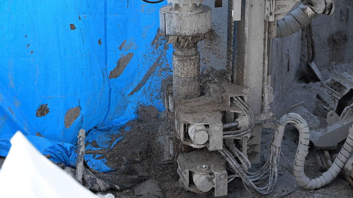 Comacchio MC 14 fully articulated microdrilling rig clamp and rotary system advancing a grout‑covered drill string against a blue tarp in a confined excavation.