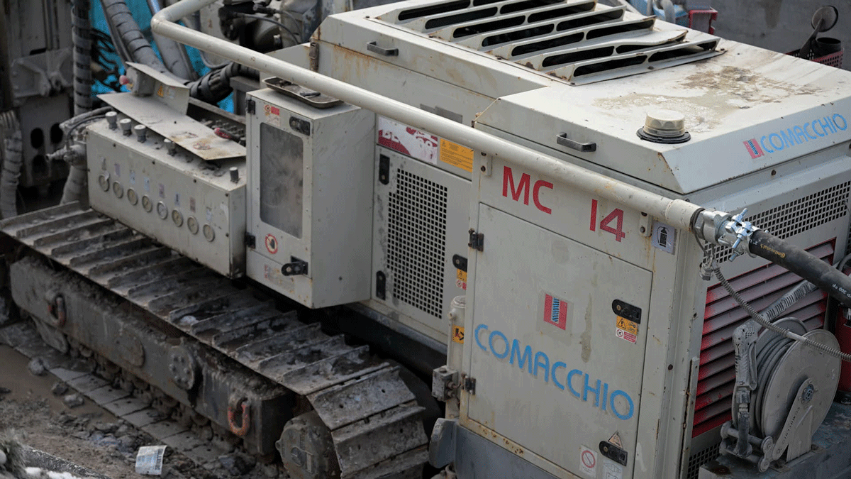 Side detail of a Comacchio MC 14 foundations and ground‑improvement microdrilling rig showing its tracked undercarriage, power pack, and control console in a muddy excavation.