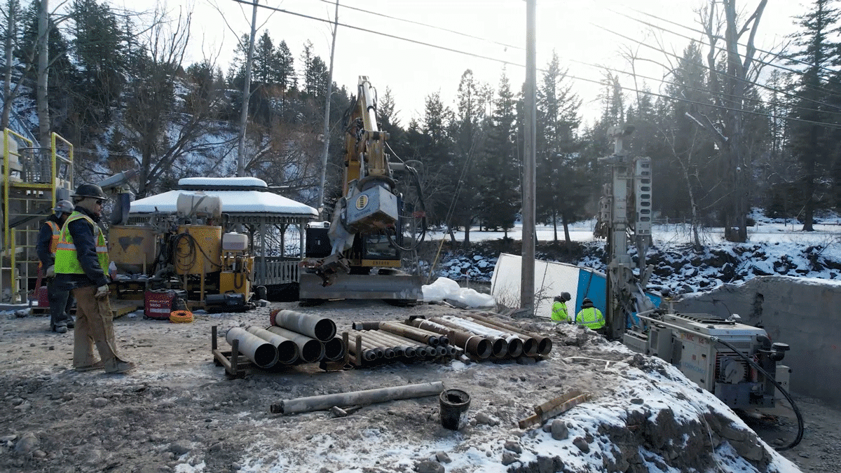 A Comacchio MC 14 foundations and ground‑improvement microdrilling rig set up in a tight, deep excavation next to a river during winter, using its compact crawler base and articulated mast to install micropiles or anchors while the crew monitors drilling progress from the slope above.