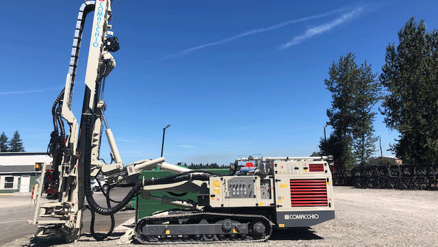 Comacchio MC 15P fully articulated multipurpose drill rig side view on tracks, mast raised, parked on a gravel yard under a clear blue sky.