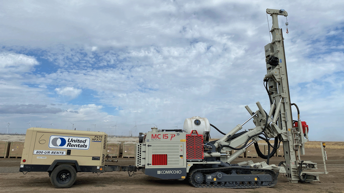 Comacchio MC 15P fully articulated multipurpose drill rig on tracks working on a wind‑farm style project site, tethered to a United Rentals air compressor under a cloudy sky.
