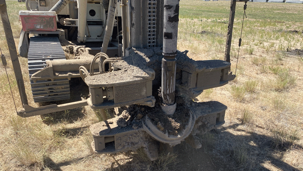 Comacchio MC 22 drill rig close‑up of the clamp and breakout system holding a mud‑covered drill string while drilling a micropile in an open field.