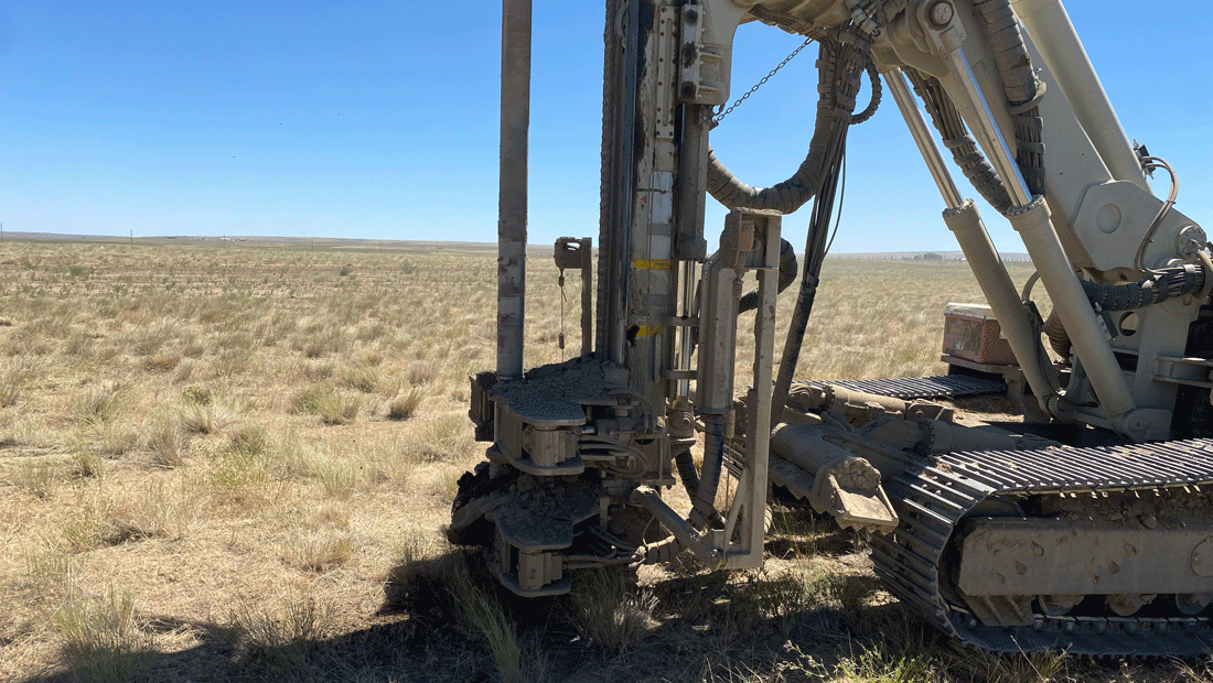 Comacchio MC 22 drill rig side view showing the mast, clamp system, and tracks while drilling a micropile in an open grassland site.