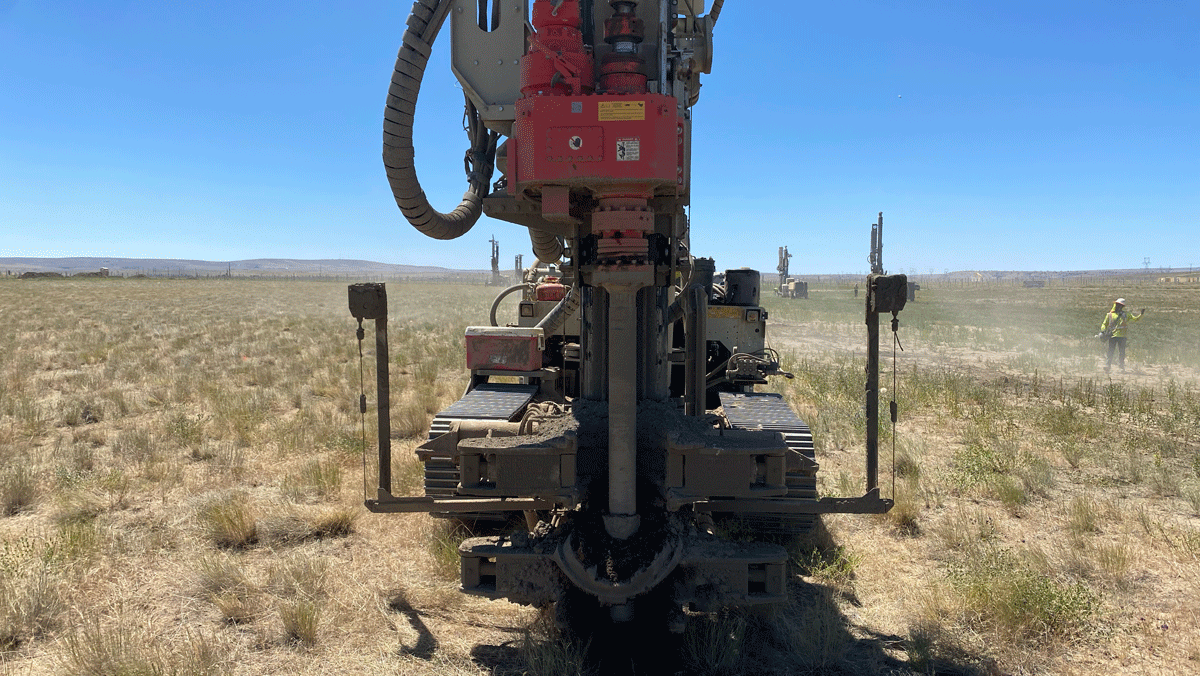 Comacchio MC 22 drill rig rear view with rotary head and mast drilling a micropile in an open field while other rigs and crew work in the background.