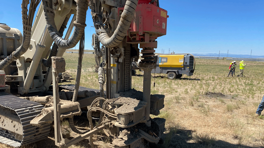 Comacchio MC 22 drill rig detail of rotary head, mast, and clamp system drilling a micropile in an open field, with compressor and crew in the background.
