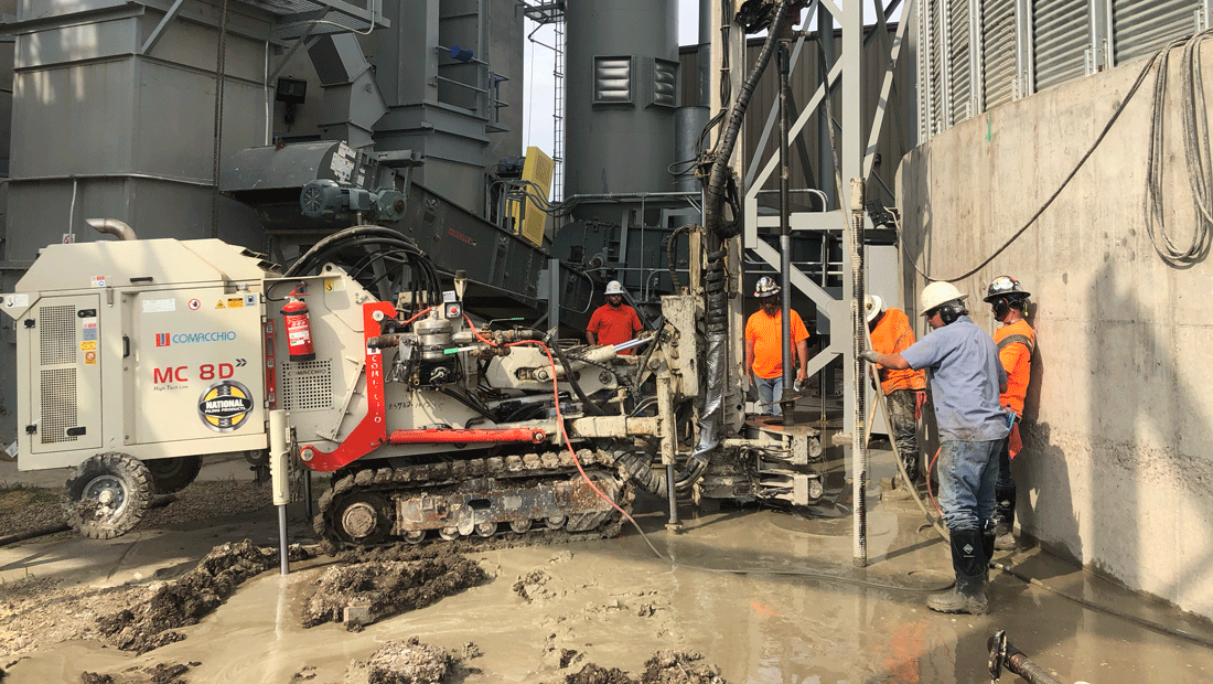Comacchio MC 8D limited‑access drill rig installing a micropile beside an industrial concrete wall, with a crew working in wet, muddy ground.