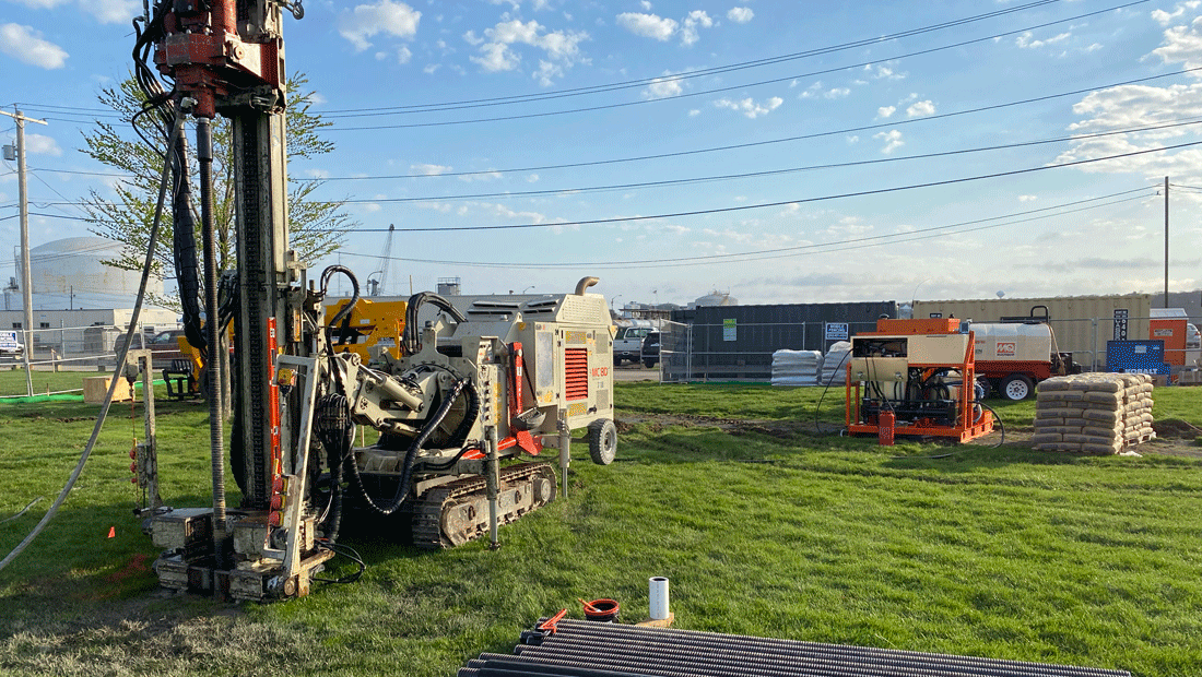 Comacchio MC 8D drill rig set up on a grassy site with steel micropile casing laid out in the foreground and a grout plant positioned in the background.