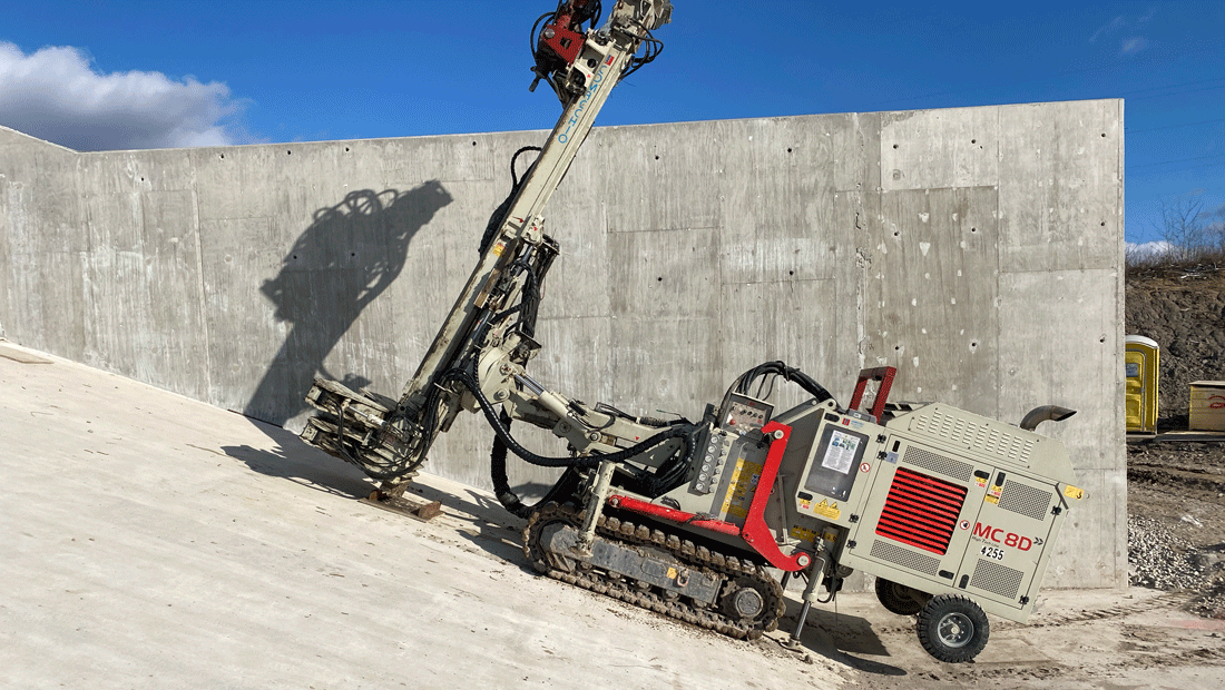 Comacchio MC 8D drill rig tracking up a steep concrete slope while positioning its mast against a tall retaining wall on a clear day.