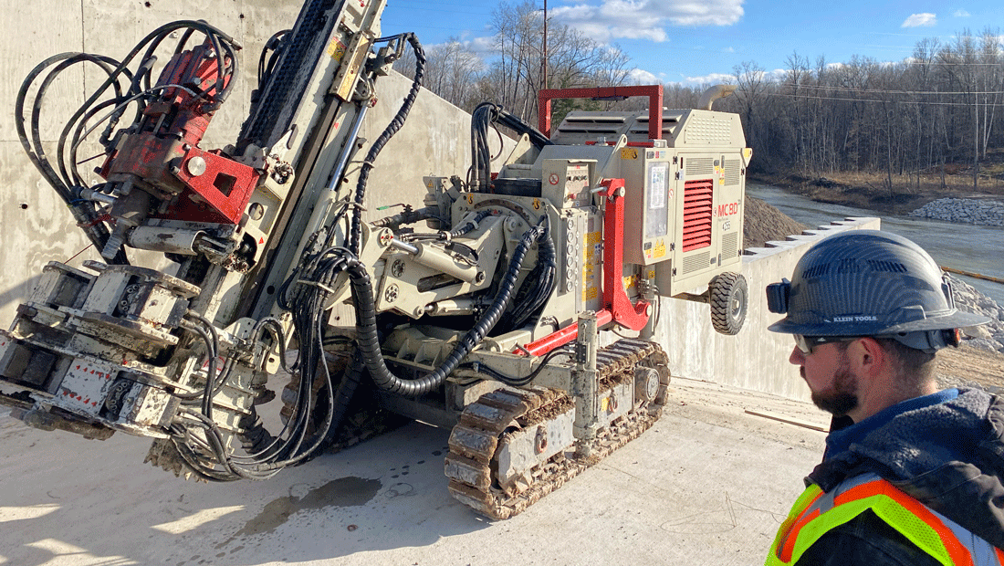 Comacchio MC 8D drill rig working on a concrete slope beside a retaining wall, with a construction worker in a safety vest and hard hat observing in the foreground.