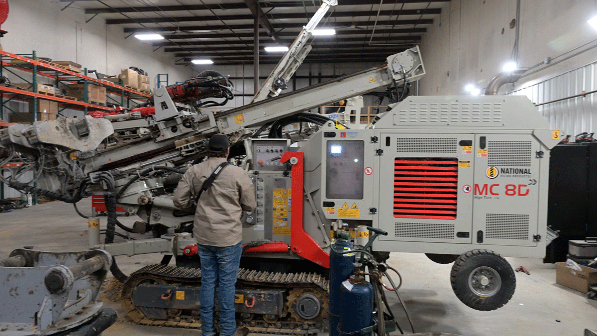 Technician standing at the control panel of a Comacchio MC 8D drill rig inside the npp maintenance shop, inspecting the machine with its mast folded.