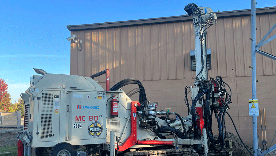 Comacchio MC 8D drill rig set up next to a metal building, drilling beside a steel tower with a casing stub visible in the dirt foreground.