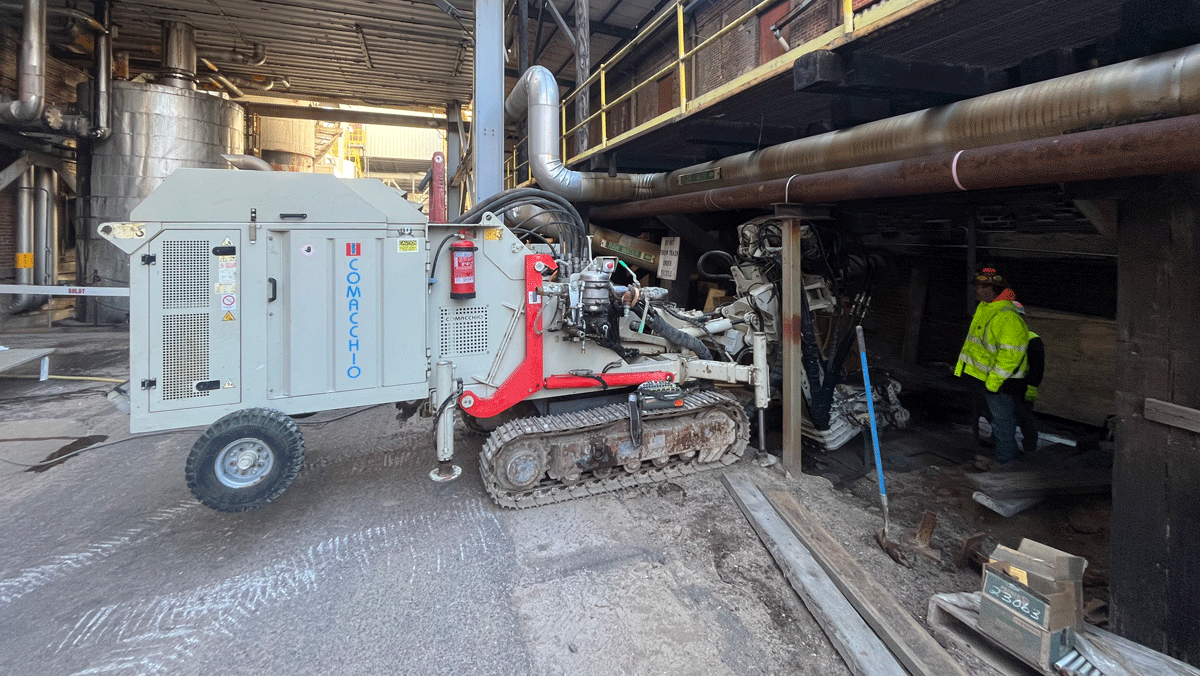 Comacchio MC 8D drill rig working under a low industrial mezzanine, installing micropiles beside a worker in high‑visibility gear.