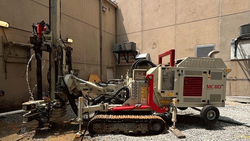 Comacchio MC 8D drill rig working in a narrow urban courtyard, drilling piles beside tall building walls with steel drill rods laid on the gravel surface.