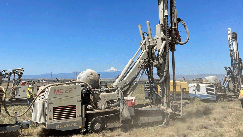 Comacchio MC 22 drill rig with tall mast raised on a remote project site, with multiple MC 22 rigs and compressors working in the background under a clear blue sky.