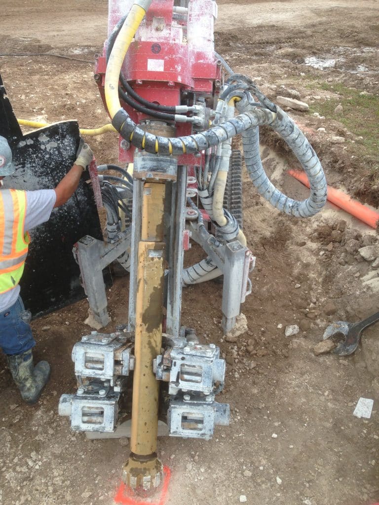 Close‑up of a down‑the‑hole hammer drill string tooling setup mounted on a foundation drilling rig, with a construction worker guiding the assembly at a marked borehole location.