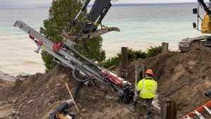 Deere excavator using a TEI HEM hydraulic excavator‑mount drilling attachment to install anchors on a steep lakeshore slope while a worker monitors drilling.