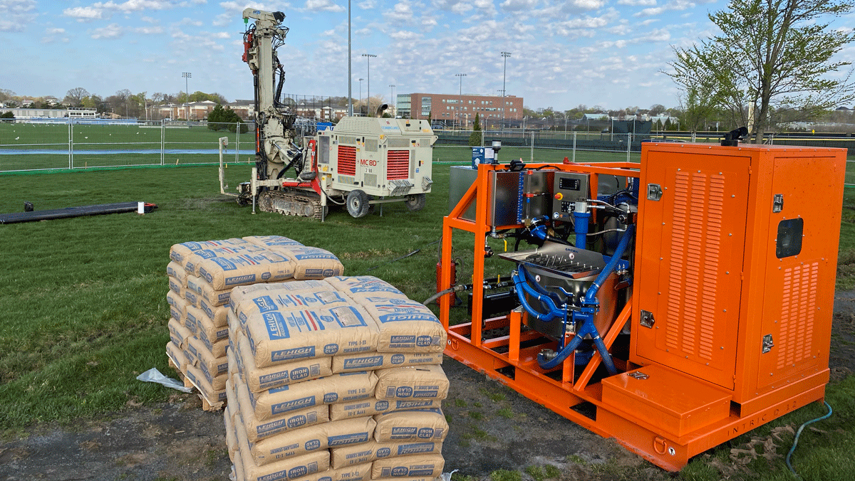 Intric D-12 high shear colloidal grout plant from National Piling Products set up on sports field project with micropile drill rig and cement bags