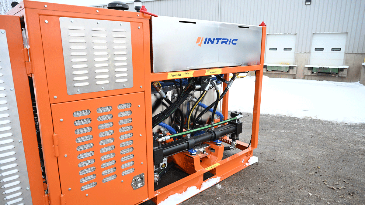 Intric D9 grout plant side view showing orange enclosure, Intric stainless tank, and exposed hydraulic components in snowy yard