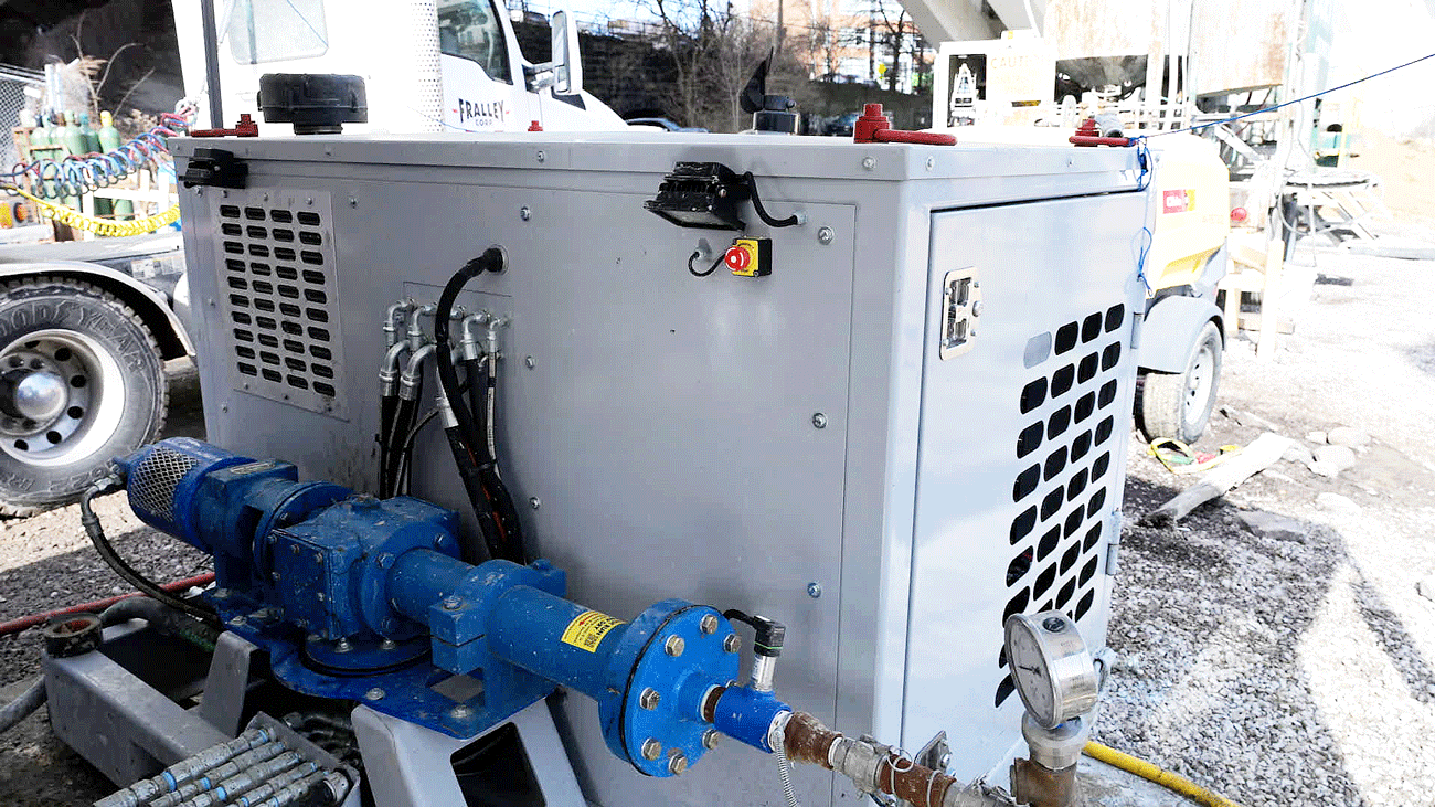 Side view of an Intric PS‑300 modular pump station on a construction site, showing the blue progressive cavity pump, hydraulic hose connections, emergency stop button, and gray enclosure with ventilation panels.