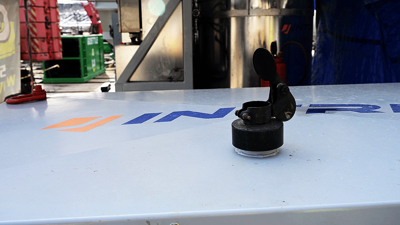 Top of an Intric PS‑300 pump station showing the black exhaust outlet with hinged rain cap and a red rigging eyelet on the enclosure roof, with construction equipment visible in the background.