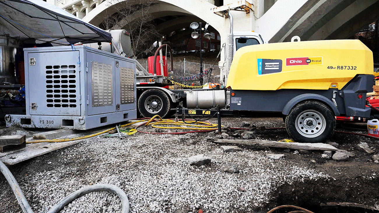 Intric PS‑300 modular pump station set up beside a yellow Atlas Copco tow‑behind air compressor on a gravel bridge repair jobsite, with hoses, cables, and a tractor‑trailer visible in the background.