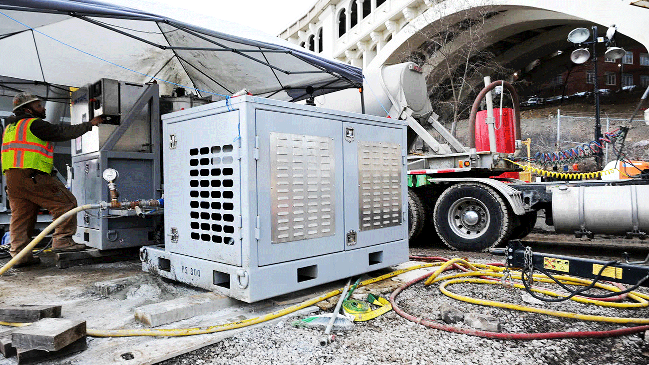 Worker in a high‑visibility vest operating grout equipment beside an Intric PS‑300 modular pump station on a bridge repair job, with the enclosed pump unit set on concrete pads and hoses running across the gravel surface.