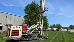 Comacchio MC-14 track drill rig set up on a manicured lawn outside the National Piling Products facility with an American flag flying from the mast on a clear day.