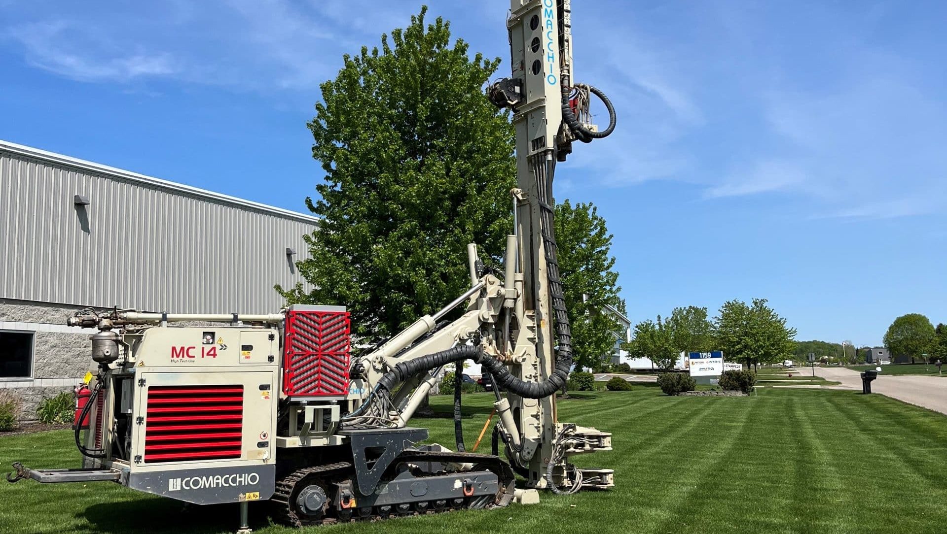 Comacchio MC-14 track drill rig set up on a manicured lawn outside the National Piling Products facility with an American flag flying from the mast on a clear day.