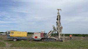 Comacchio MC15P track drill rig working in an open field while connected to a trailer‑mounted generator and monitored by a construction worker.