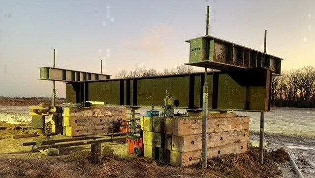 Steel National Piling Products test beam setup on stacked concrete blocks and threaded reaction posts at a jobsite, with hydraulic jacks and hoses arranged below for load testing.