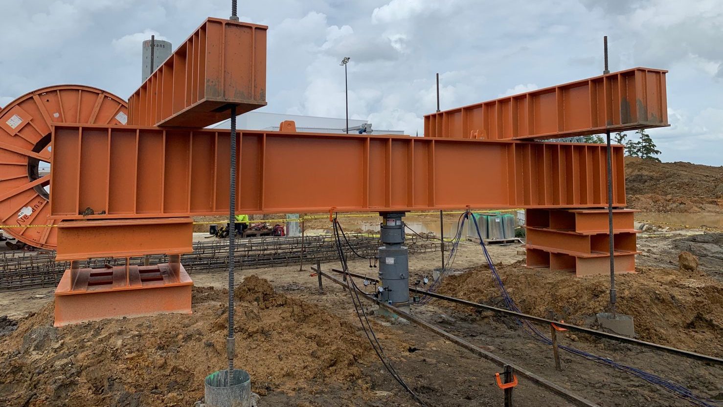 Large orange National Piling Products test beam frame assembled on threaded reaction posts above a pile head, with hydraulic jack and hoses set up for high‑capacity load testing on a construction site.