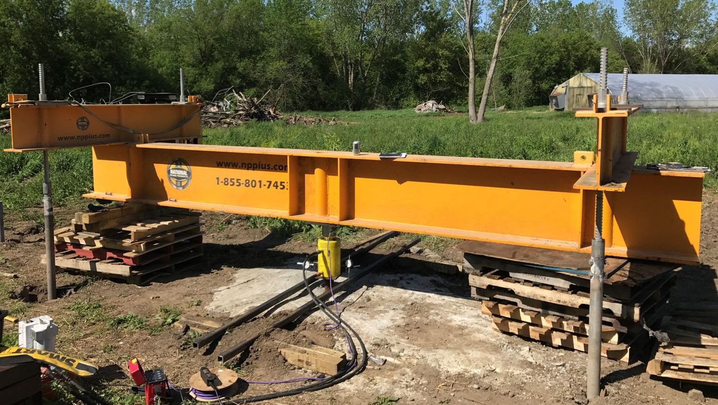 Yellow National Piling Products test beam set up outdoors on screw jacks and timber cribbing, with hydraulic jacks and hoses connected for tension and compression load testing of deep foundations.