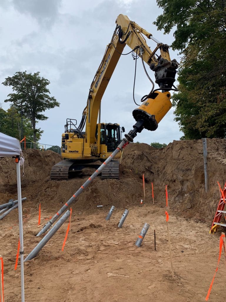 Komatsu excavator using a ProDig 60K excavator‑mounted screw pile driver to install battered piles on a deep excavation project.