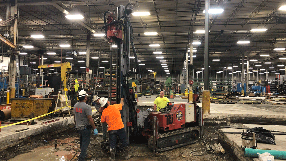 TEI TD‑100 electric‑hydraulic limited‑access drill rig installing interior micropiles with crew gathered around the mast in a large industrial plant.