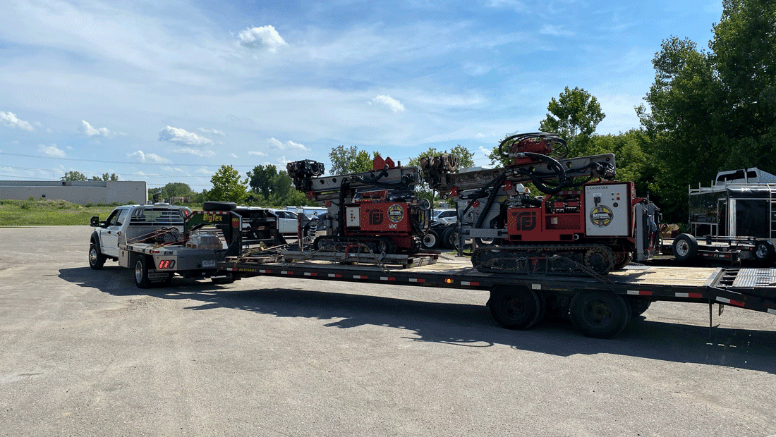 Truck hauling two TEI TD‑75 electric‑hydraulic limited‑access drill rigs on a gooseneck trailer from National Piling Products yard.