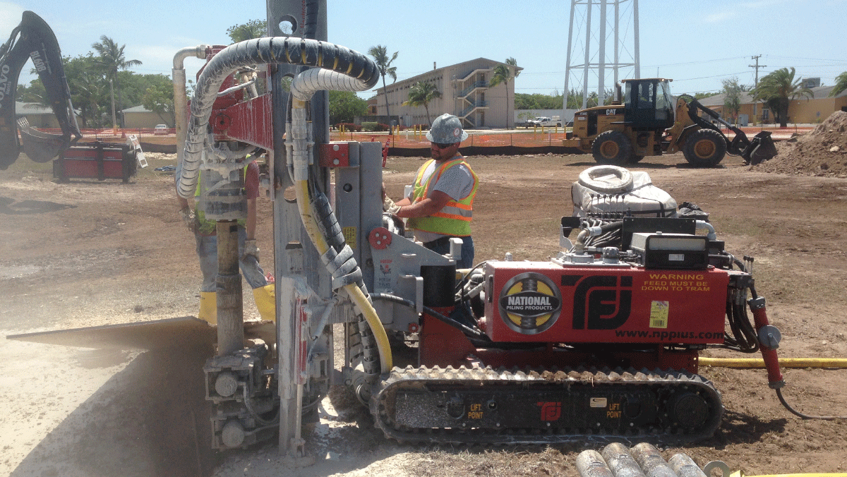 TEI TD‑75 electric‑hydraulic limited‑access drill rig installing micropiles on an open jobsite with National Piling Products crew and earthmoving equipment in the background.