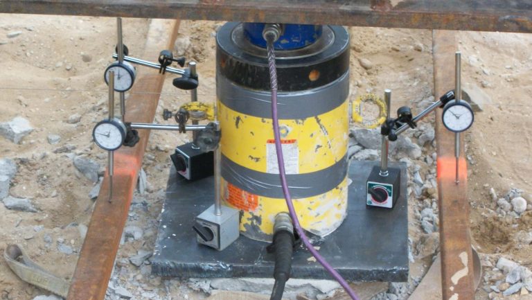 Hydraulic jack and dial gauge setup performing a static load test on a deep foundation pile with indicators mounted on steel beams to measure movement.
