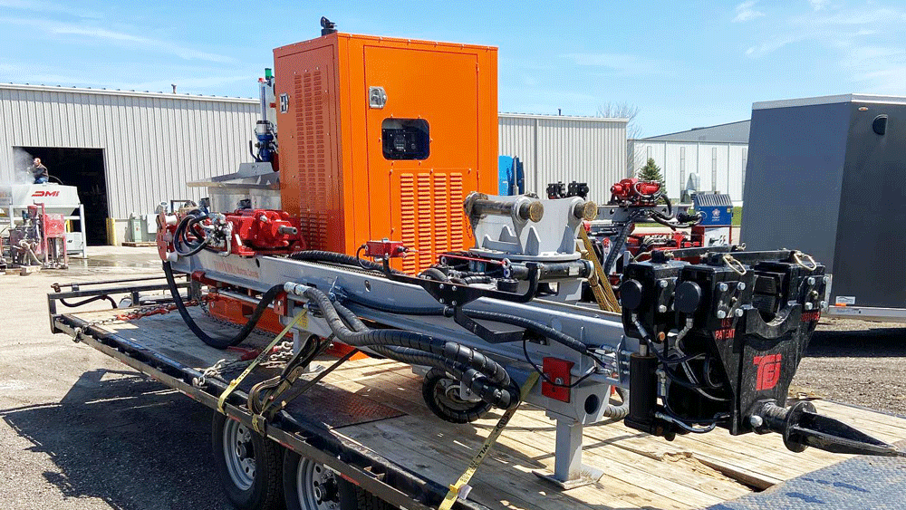 TEI HEM 760 excavator‑mounted drilling attachment strapped to a flatbed trailer, showing the grey feed boom, black TEI drifter, hydraulic hoses, and orange power pack in an industrial yard.
