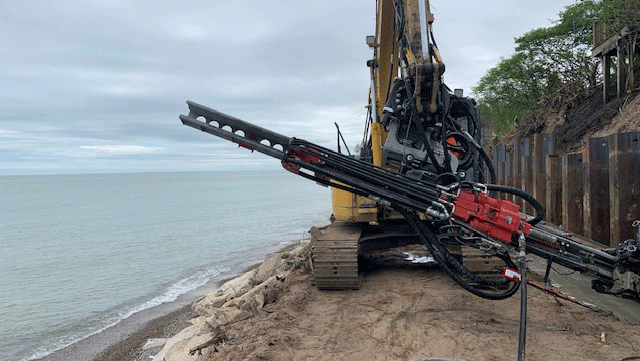 National Piling Products rental TEI HEM hydraulic excavator‑mounted drilling attachment working along a steel seawall to stabilize a sandy shoreline.