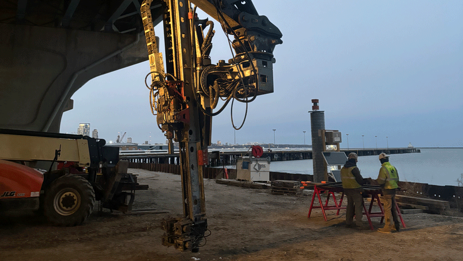 National Piling Products TEI HEM hydraulic excavator‑mounted drilling attachment working under a bridge along the waterfront, with two crew members reviewing plans nearby.