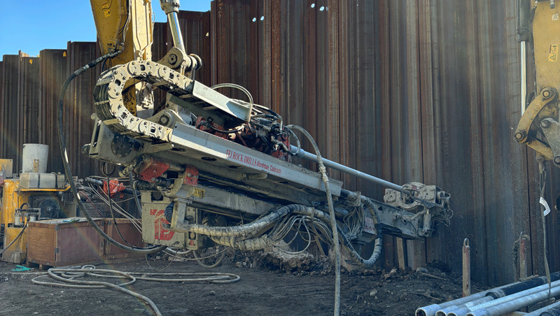 National Piling Products TEI HEM hydraulic excavator‑mounted drilling attachment installing anchors along a steel sheet‑pile wall, with drill rods staged nearby.