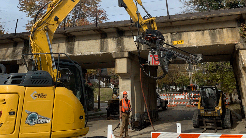 National Piling Products TEI MME hydraulic mini‑excavator drilling attachment mounted on a Kobelco excavator installing anchors beneath a low railroad bridge in a city street work zone.